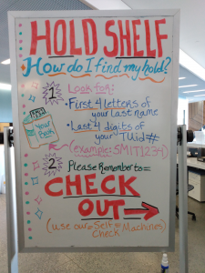 Whiteboard sign next to the One Stop desk that explains to users how to locate and checkout their items on the hold shelf
