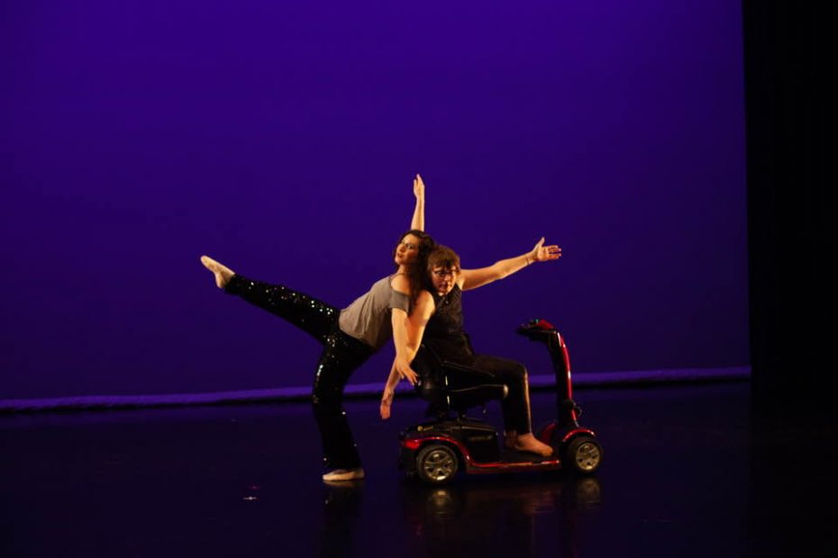 Two dancers are “fused” back to back. One stands with a leg extended in the air. The other is seated on a red mobility scooter with both of her arms extended and back slightly arched.