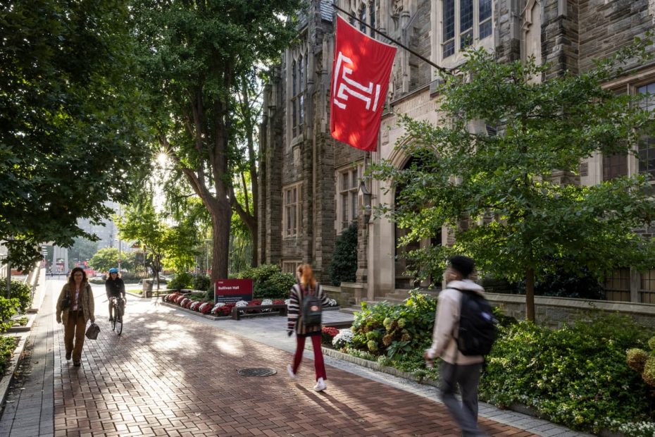 Polett Walk on Temple's campus features stone buildings, large red and white Temple T flag, green plants and flower, and students walking and cycling