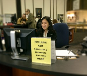 Girl sitting at reference desk behind sign reading: Tech Help, Ask Computer and Technical Questions Here.