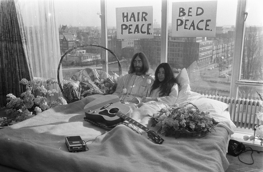Famous black and white image of John Lennon and Yoko Ono in bed. A guitar, flowers, and a tape recorder sit on the bed with them.
