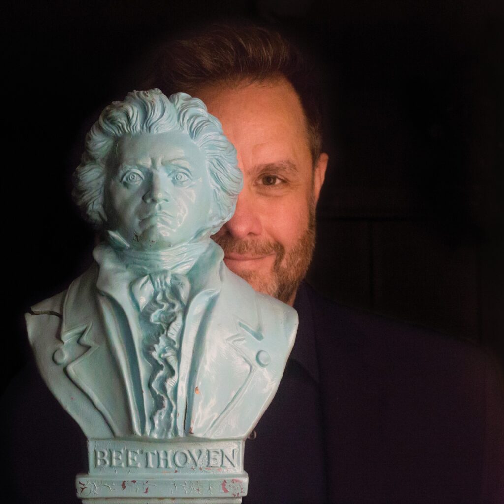 Photograph a bust of Beethoven, half obscuring the smiling face of a white man with facial hair.