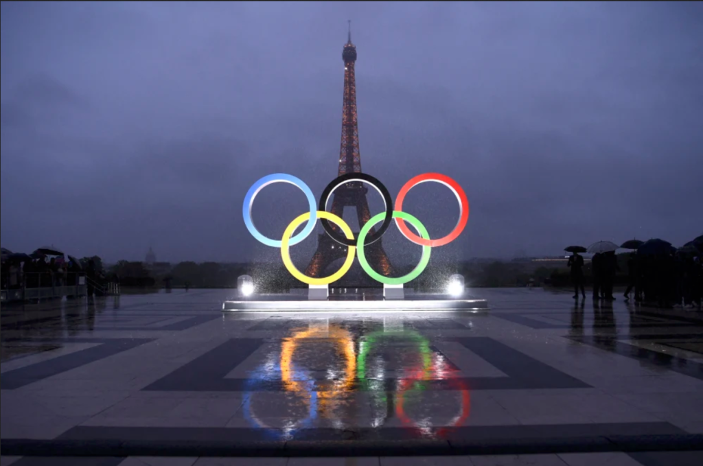 Photograph of the Olympic Rings with the Eiffel Tower in the background and small grounds of people carrying umbrellas on either side.