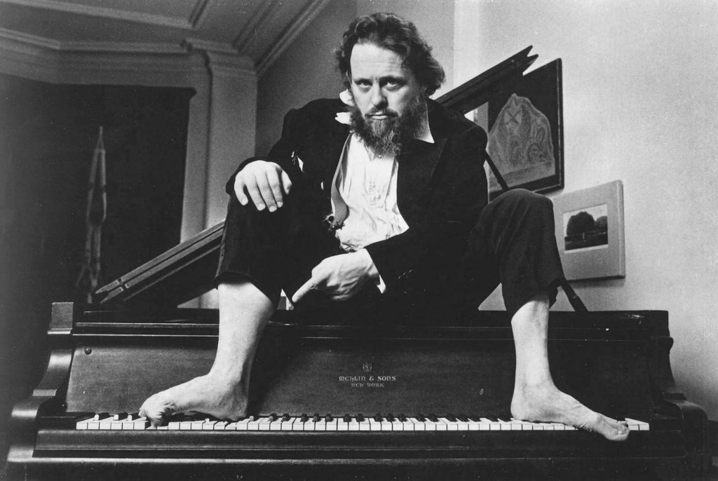 Black and white photograph of a barefoot man in concert attire sitting on a piano with his feet splayed on the keys.