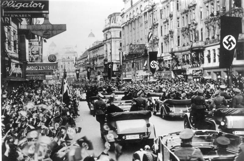 Black and white photograph of a busy city street filled with old-fashioned cars in bumper to bumper traffic. A flag with a swastika looms prominently in the corner.