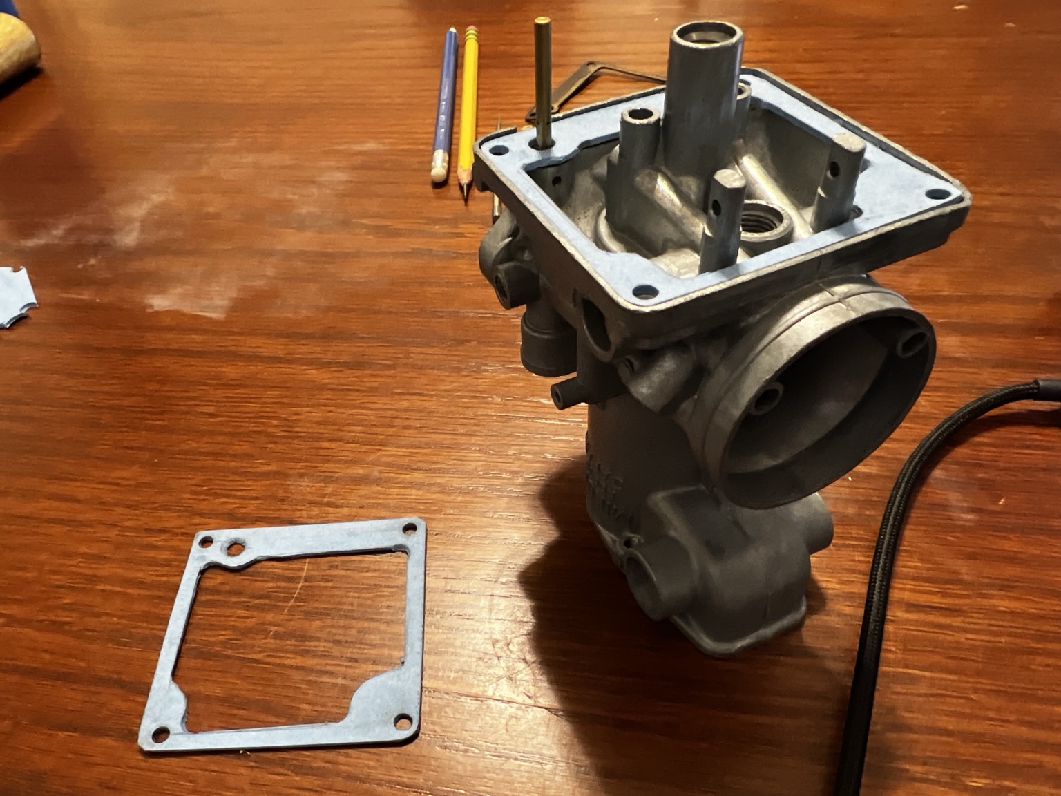 Hand-cut blue gaskets beside and fitted to a motorcycle carburetor on a wooden work surface.