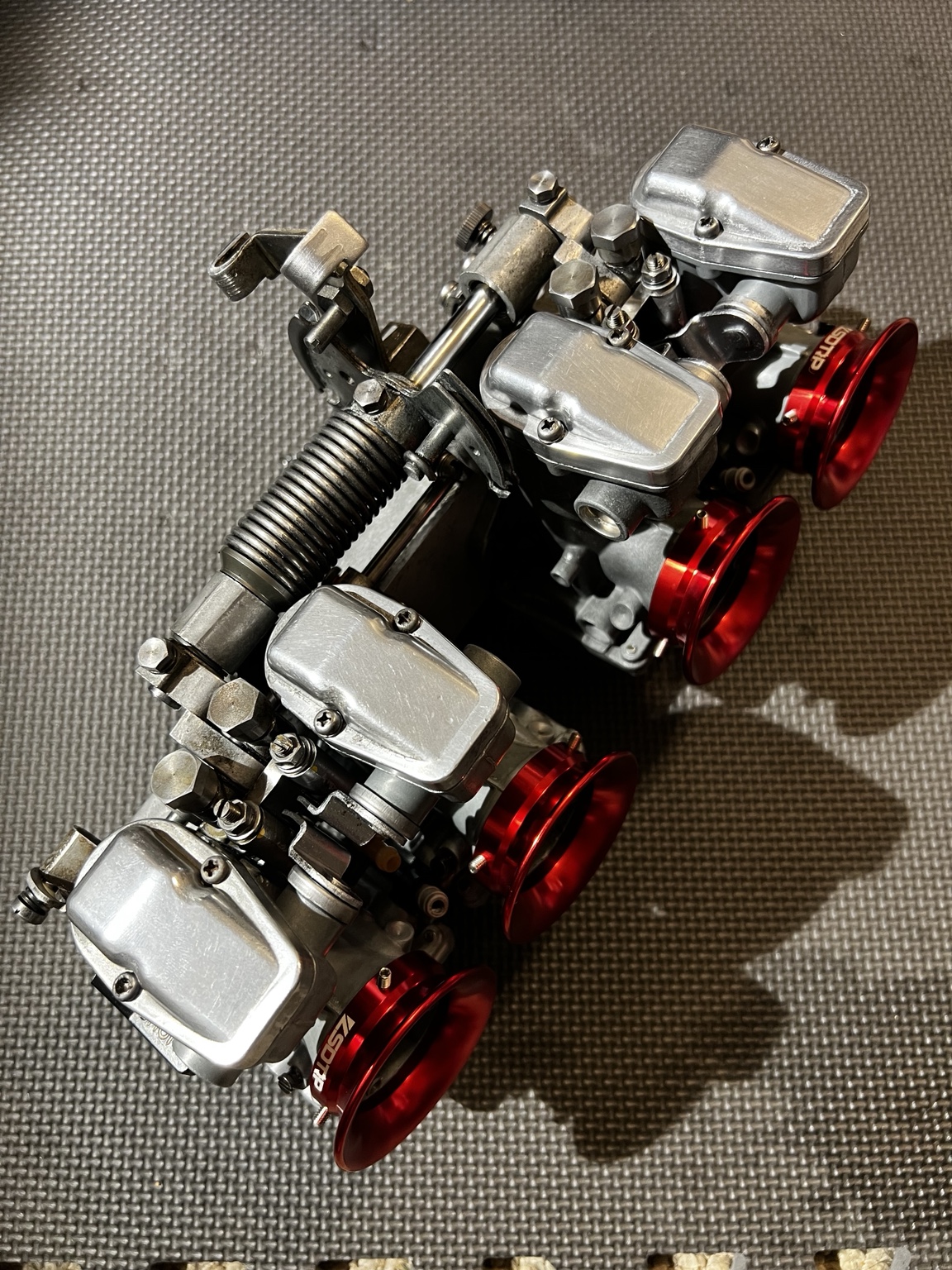 A bank of four assembled motorcycle carburetors fitted with red velocity stacks.