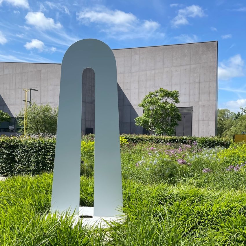 A painted steel sculpture by Kim Lim stands outdoors in a lush garden at the Hepworth Wakefield, with the museum's modern concrete facade visible in the background under a bright blue sky with scattered clouds. The abstract artwork consists of a tall, arch-like form with an elongated oval cutout in the center.