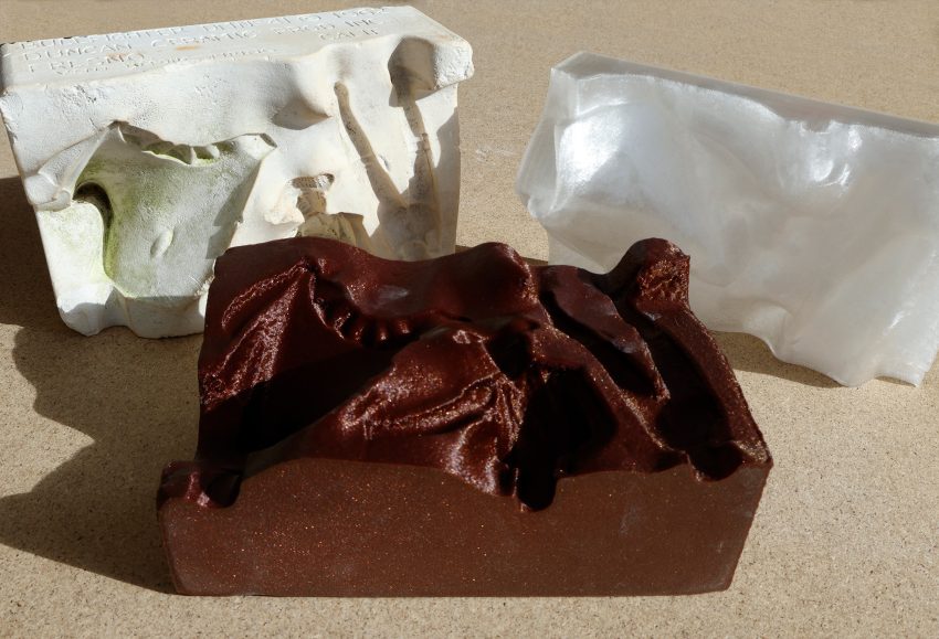 Right: half of a plaster mold for a matador figurine sitting upright. Text on top of the mold indicates it was produced in Fresno for school use. Middle: a copy of the mold 3D printed from a scan in sparkly brown filament laying flat on on MDF table. Right: the inverse of the mold printed in clear filament sitting upright.