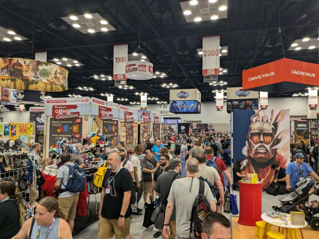 A crowd of people make their way between the booths that make up one aisle of the exhibit hall at Gen Con.